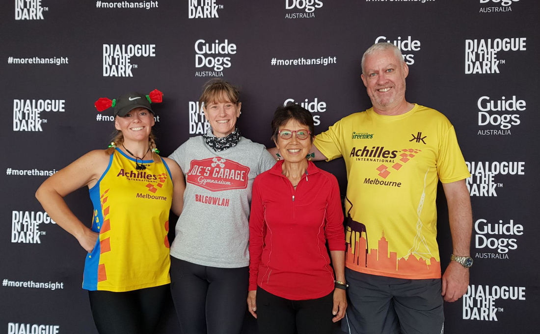 Four Achilles guides stand arm in arm in front of a black Dialogue in the Dark and Guide Dogs Australia banner