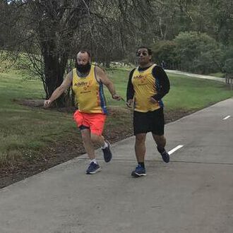 Paul, left, wearing Achilles singlet and orange shorts guides Amir via a hand-hand tether. Amir wears an Achilles singlet and black shorts. They are running on a concrete path with a weeping winter tree behind them