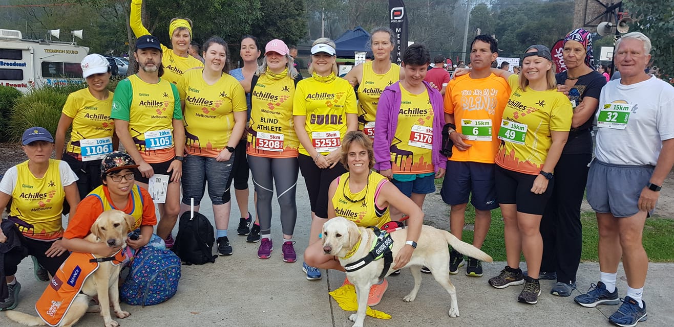 A group photo of around 16 Achilles athletes standing side by side, two squatting at the front with two guide dogs. 