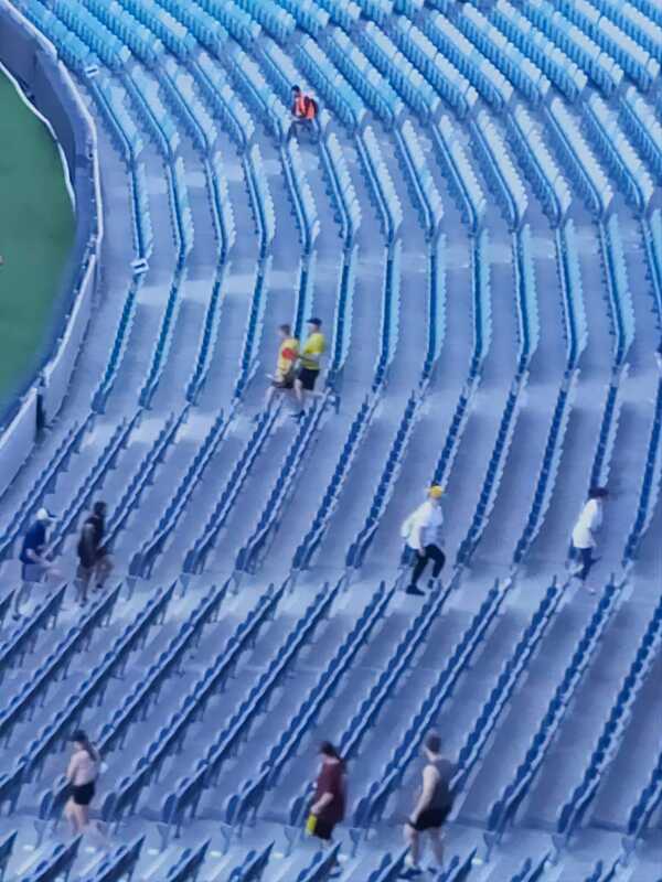 A far away shot of Francois and Simon stomping down the MCG stadium stairs on the level 1 stands. They are two dotted yellow-glad people in amongst rows and rows of empty chairs with other stompers climbing stairs in the adjacent aisles
