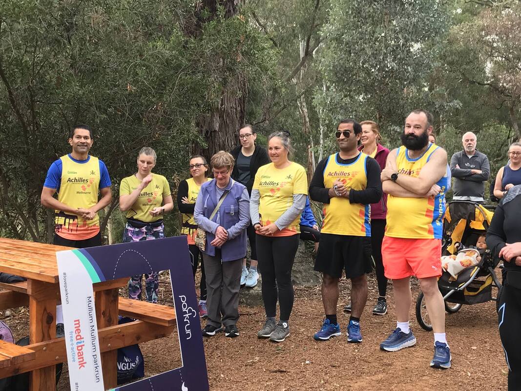 Group of Achilleans stand side by side listening to a speech, Paul stands to the right of the group with arms crossed listening intently. Parkrun cardboard social media frame in foreground and other parkrunners with trees in background. 