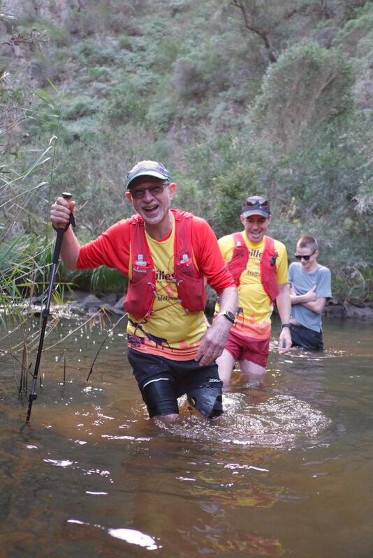Peter in the foreground wears a red vest over his Achilles top and holds a walking pole planted into the river on his right. He has a big smile on his face and is thigh-deep in water. Jez walks behind him, also thigh-deep in water, and his son Aiden behind him with his hands tucked into his armpits. Shrubs on the river bank extend up the hill behind them.
