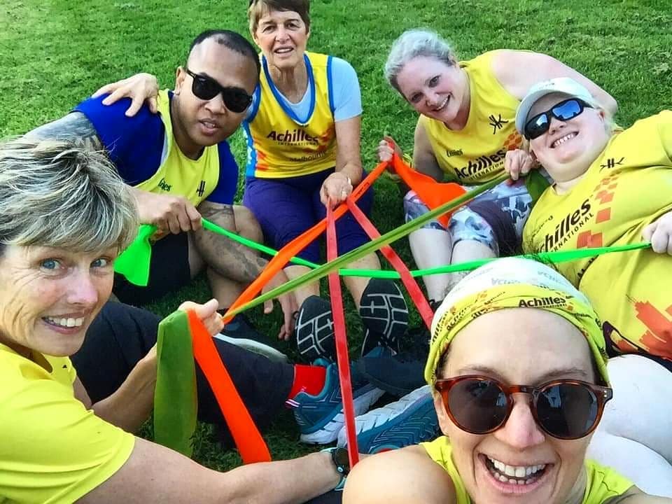 A selfie with 6 people sitting on grass, Sarah with sunglasses and an Achilles buff is in the bottom right of the camera. The other 5 people are arranged in a seated circle with a tangled arrangement of colourful exercise bands between them. 