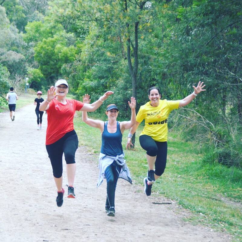 Three ladies (left to right) Sonia in red, Wendy in blue and Julie in Achilles yellow are jumping in the air in the middle of their run. Gravel track extends behind them and shrubs and trees are seen bordering the track.