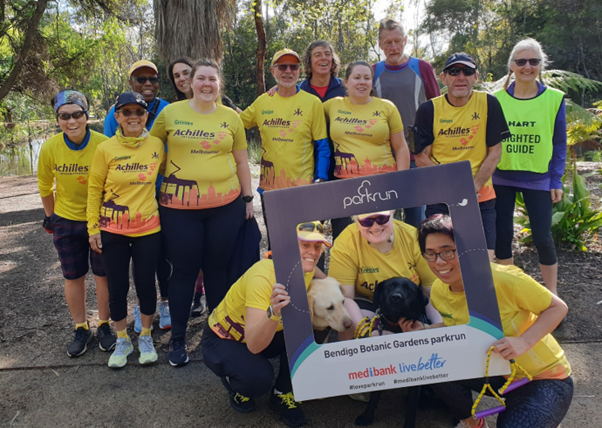 A group of 11 Achilles athletes and Bendigo parkrun crew with three more athletes and 2 dog guides squatting below holding the parkrun social media frame that says 