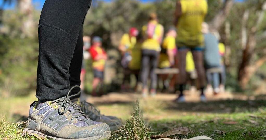 Close up photo of grey-blue hiking shoes and lower legs pictured up to the shin in foreground, blurred Achilleans in yellow shirts and hiking packs gathered in a group in the background and out of focus.