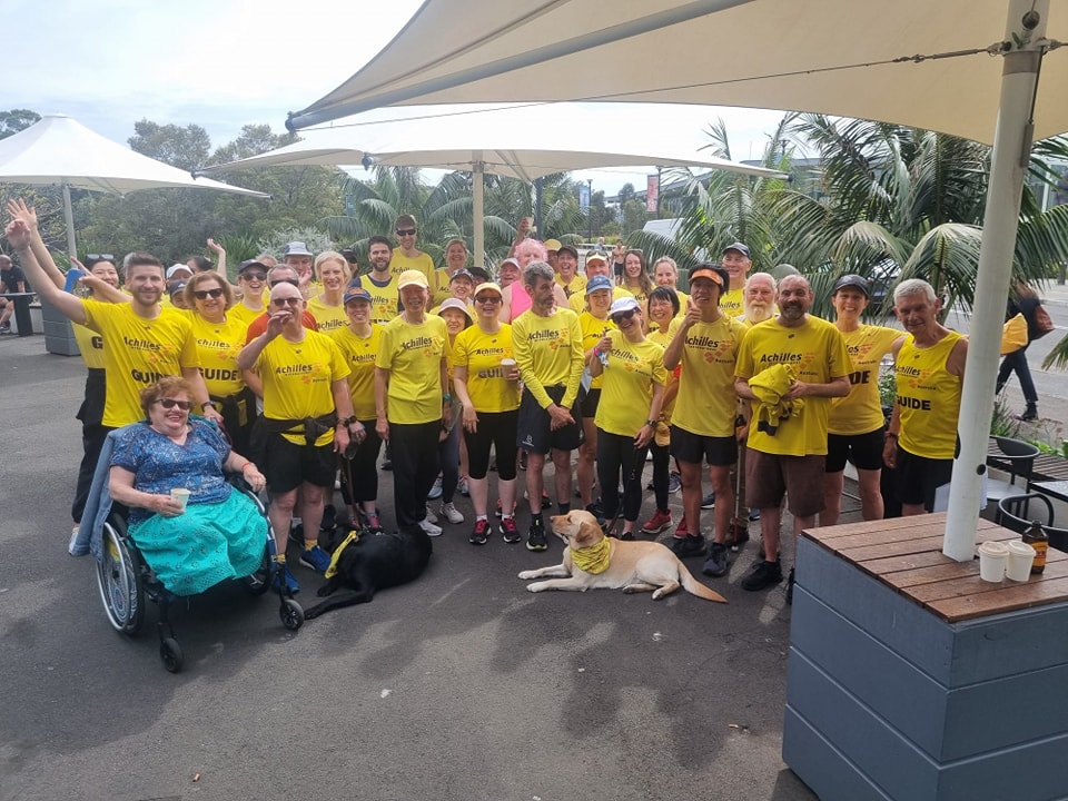 A large group photo of approximately thirty five Achilles team members standing under large shade cloths smiling post coffee with two guide dogs