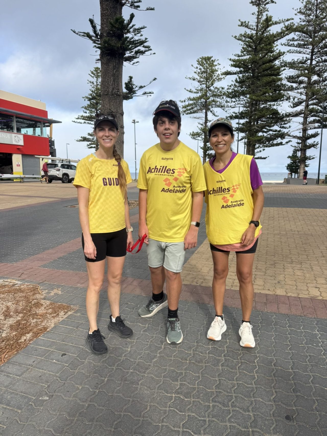 From left to right, Danielle, Alex and Paola standing together at the Glenelg Rotunda in March 2026. They are wearing yellow Achilles Adelaide tops and are smiling. 